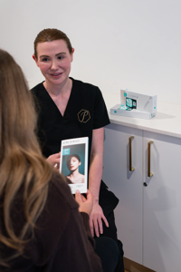 Clinician in black uniform consulting with client holding a skincare brochure in a clinic room