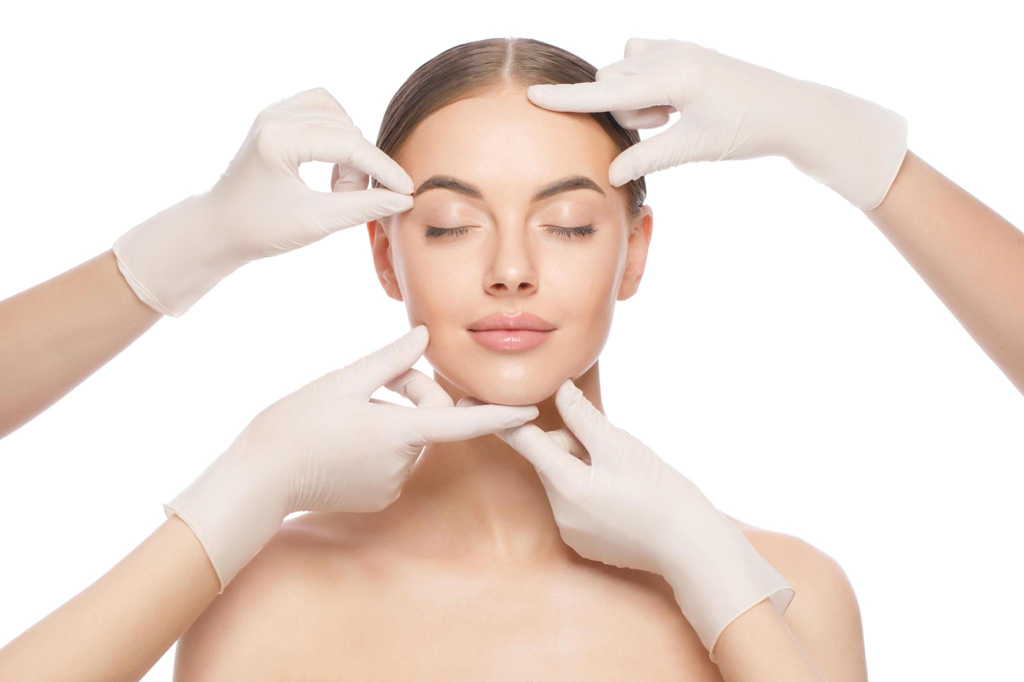 Woman with closed eyes having her face examined by hands in white gloves on white background