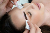 Close-up of woman receiving eyebrow treatment with hands applying product and wiping brow area