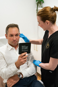 Man in white shirt holding a mirror while clinician in black scrubs with blue gloves examines his face